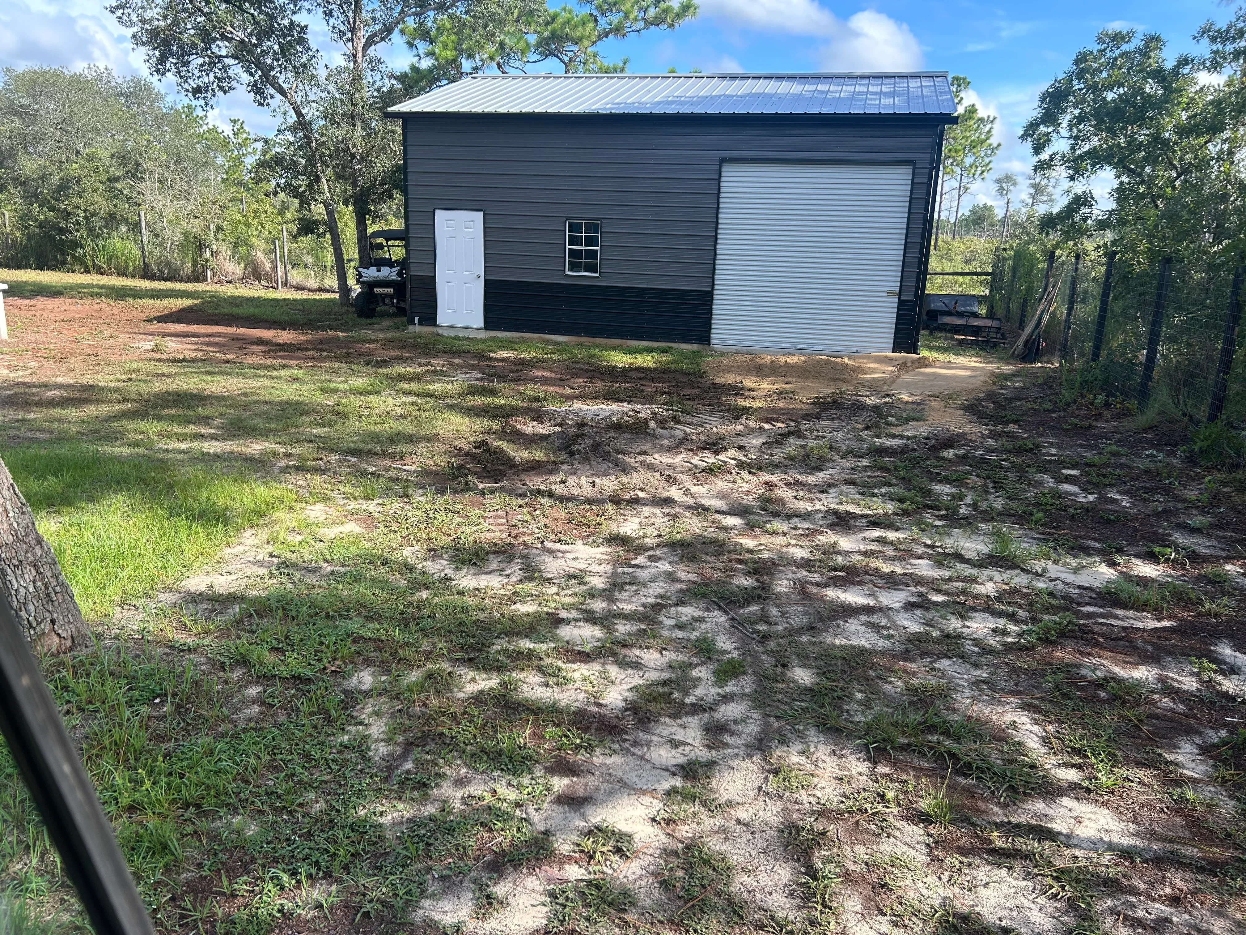 Black metal building with white door and garage door on graded dirt lot surrounded by trees