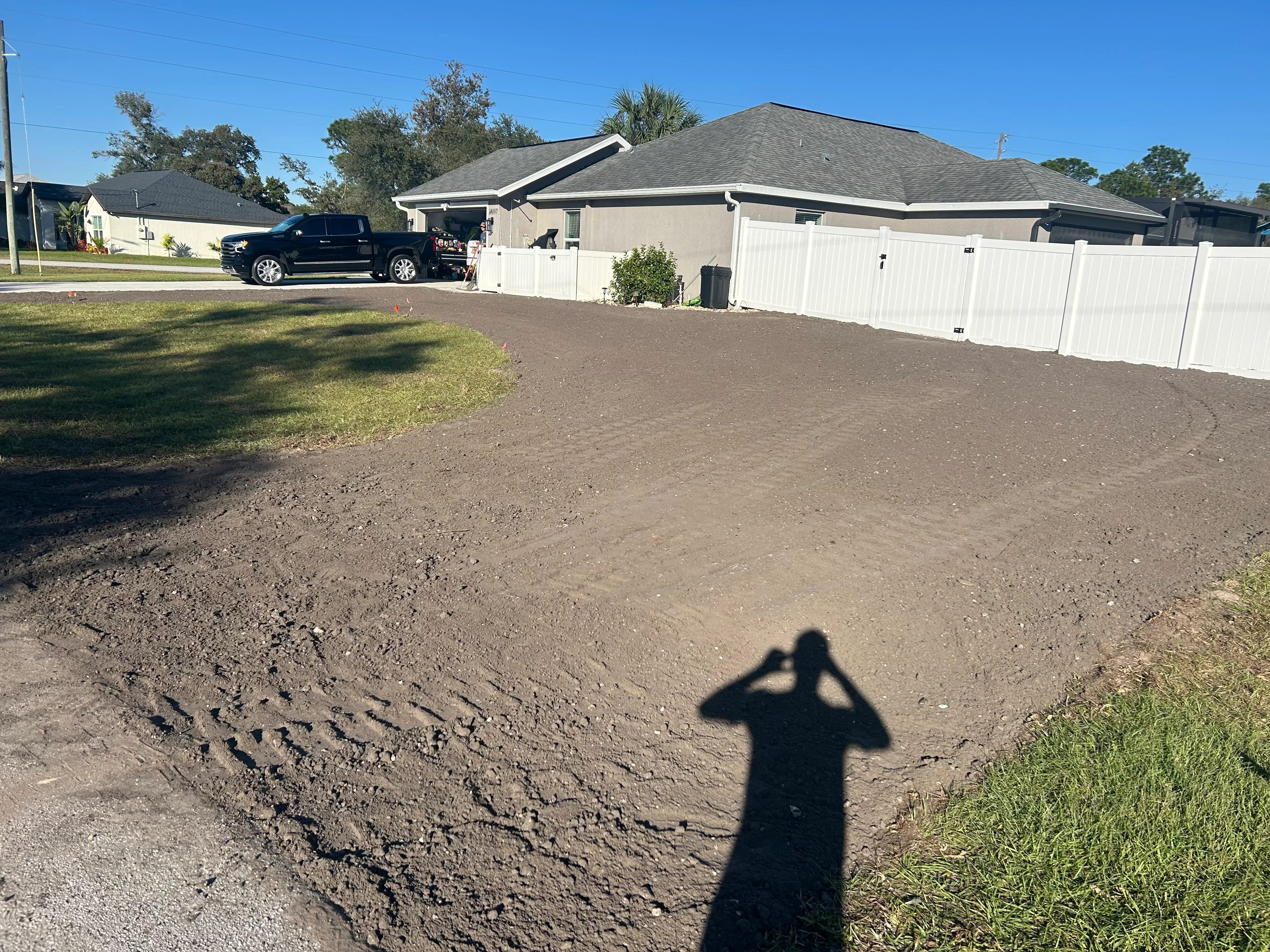 Excavated and graded driveway base with compacted soil, white vinyl fence, and residential home