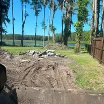 Excavated residential lot with exposed soil, cleared debris pile, and mature trees along property edge