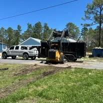 Track loader and white pickup truck on grass lot with pine trees in background