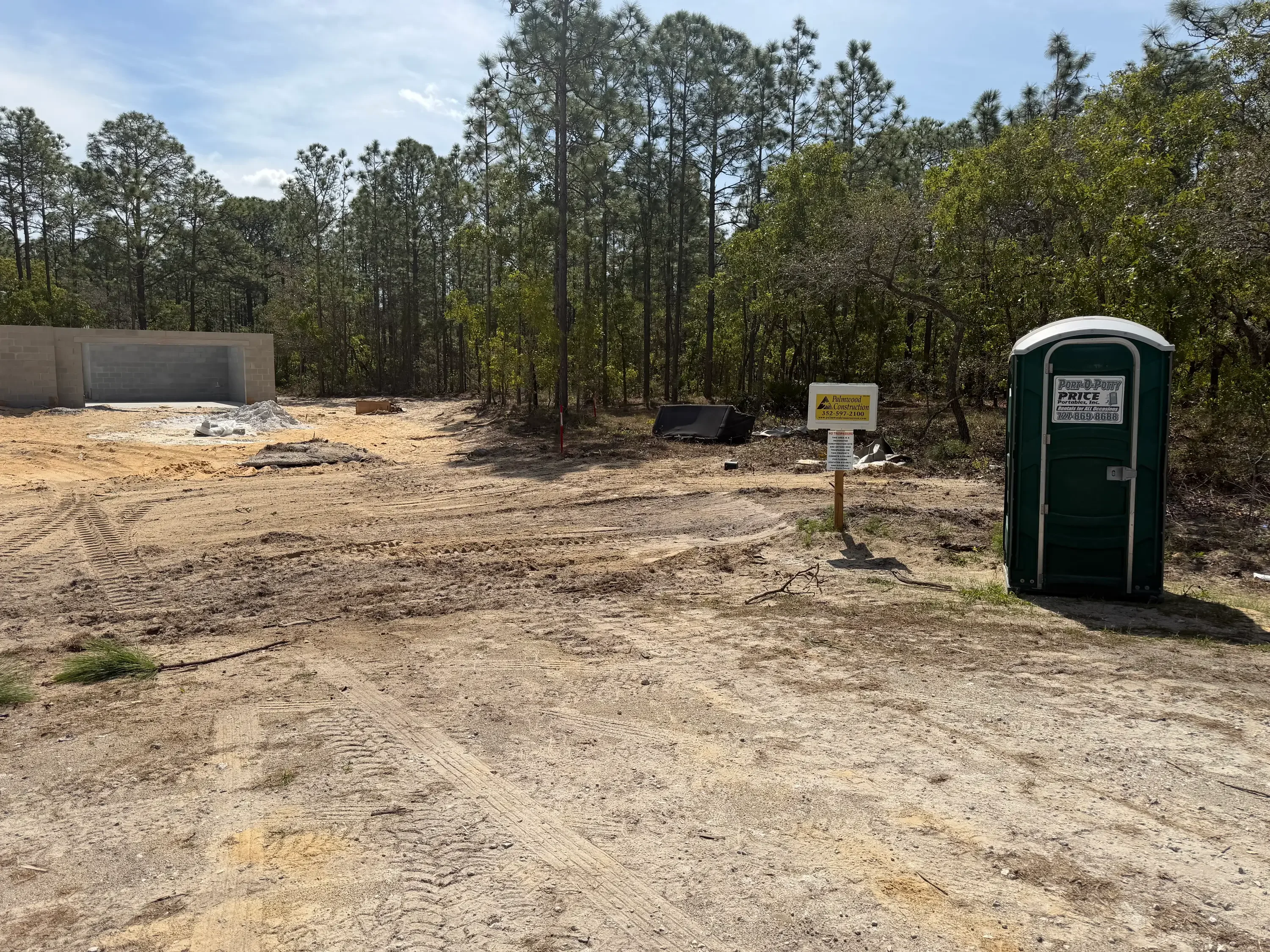Excavated construction site with graded dirt, concrete foundation pad, and portable facilities among pine trees